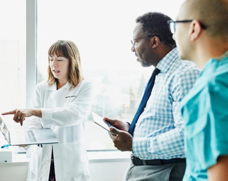 Female doctor holding laptop discussing patient information with medical team in exam room - stock photo
