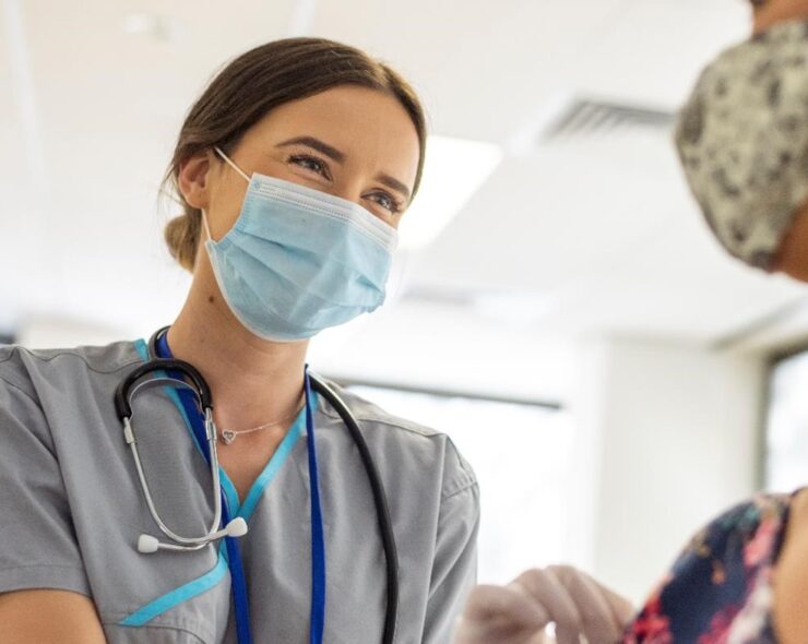 Shot focused on a kind looking nurse wearing scrubs, a stethoscope, white rubber gloves and a protective face mask. She is smiling at the nervous patient with her eyes.