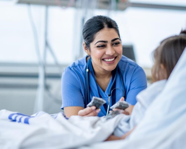 A female nurse of Middle Eastern decent sits at the edge of a hospital bed as she check in on her young patient. She is wearing blue scrubs and is attempting to cheer the young girl up.