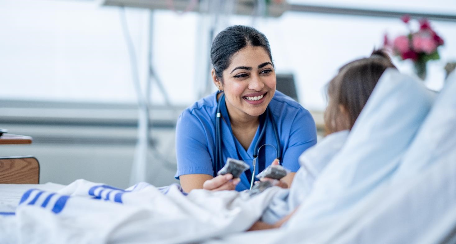 A female nurse of Middle Eastern decent sits at the edge of a hospital bed as she check in on her young patient. She is wearing blue scrubs and is attempting to cheer the young girl up.